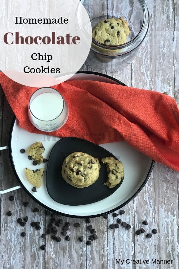 In the background is a jar with cookies in it. There is a white tray that has an orange napkin on it with a grey plate that has chocolate chip cookies on it. There is also a glass with milk on the tray. In front of the tray is chocolate chips.
