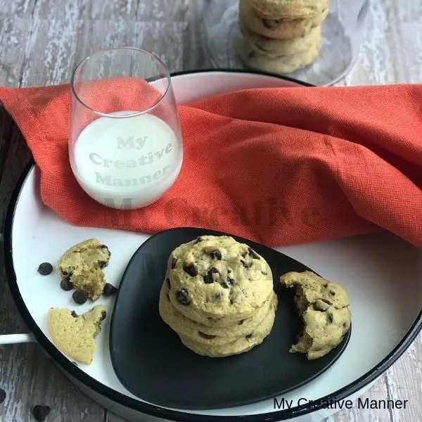 In the background is a jar with the best ever chocolate chip cookies in it. There is a white tray that has an orange napkin on it with a grey plate that has chocolate chip cookies on it. There is also a glass with milk on the tray. In front of the tray is chocolate chips.