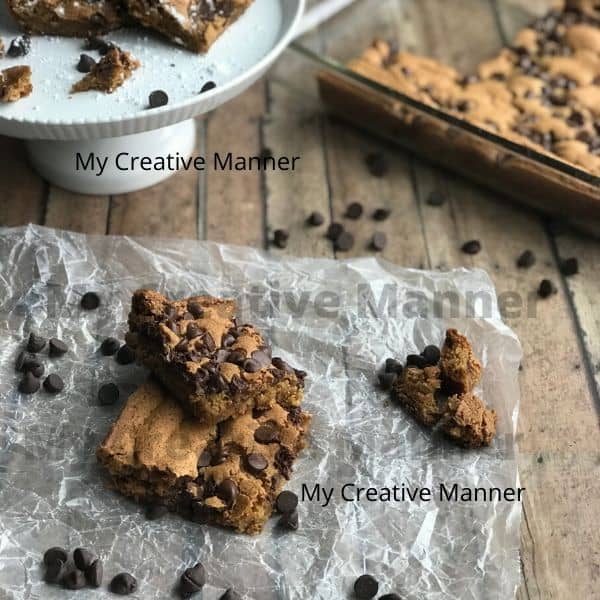 Three blonde brownies on wax paper with chocolate chips around them. A white cake stand that has more blonde brownies on it. A baking dish the more blonde brownies in it.