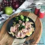 Grey and white towel with grapes and an apple on it with a plant next to that. Red salt and pepper shakers are next to a wooden board that has a plate with beef stroganoff and broccoli on it.
