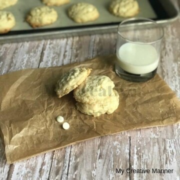 Cookie sheet in the background with cookies on it. Amazing White Chocolate Coconut Cookies on a brown paper with a glass of milk next to it.
