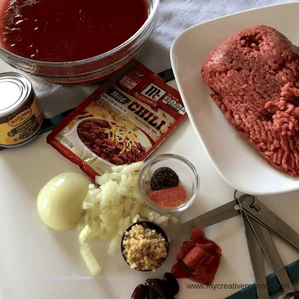 All the ingredients to make chili; ground beef, tomato sauce, onion, seasonings, garlic, tomato paste, and beans.