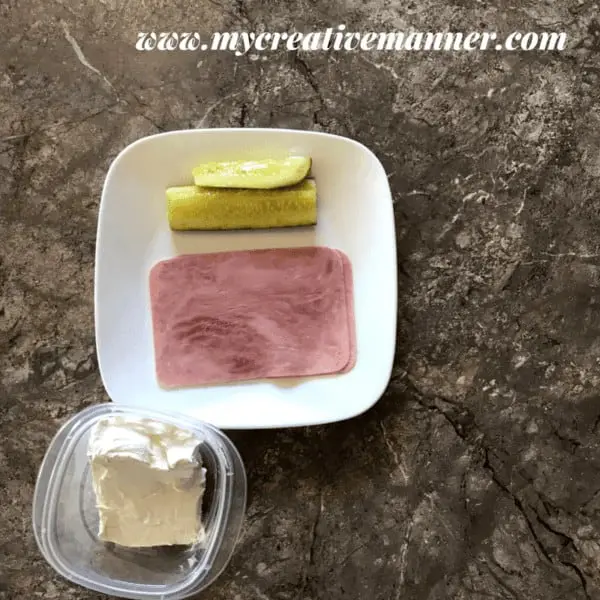 White plate that has a slice of ham and pickle on it. A bowl that has cream cheese in it.