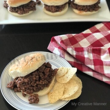 Three sloppy Joes on a white plate in the background. A red and white checker napkin is behind a white plate that has a sloppy Joe on it with chips.