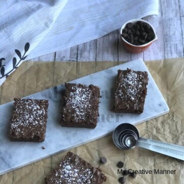 Three Killer Fudge Brownies on a plate with measuring spoons in front of the plate. On the brown paper is another killer fudge brownie and chocolate chips. Behind the plate is a measuring cup full of chocolate chips and a towel.