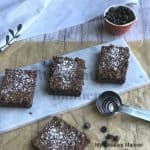 Three Killer Fudge Brownies on a plate with measuring spoons in front of the plate. On the brown paper is another killer fudge brownie and chocolate chips. Behind the plate is a measuring cup full of chocolate chips and a towel.
