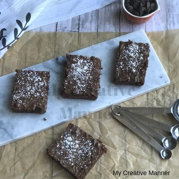 Three Killer Fudge Brownies on a plate with measuring spoons in front of the plate. On the brown paper is another killer fudge brownie and chocolate chips. Behind the plate is a measuring cup full of chocolate chips and a towel.