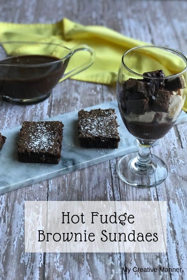 Hot fudge sauce in a gravy boat with a yellow napkin in the background. Three brownies on a plate and a glass that is filled with the Hot Fudge Brownie Recipe.