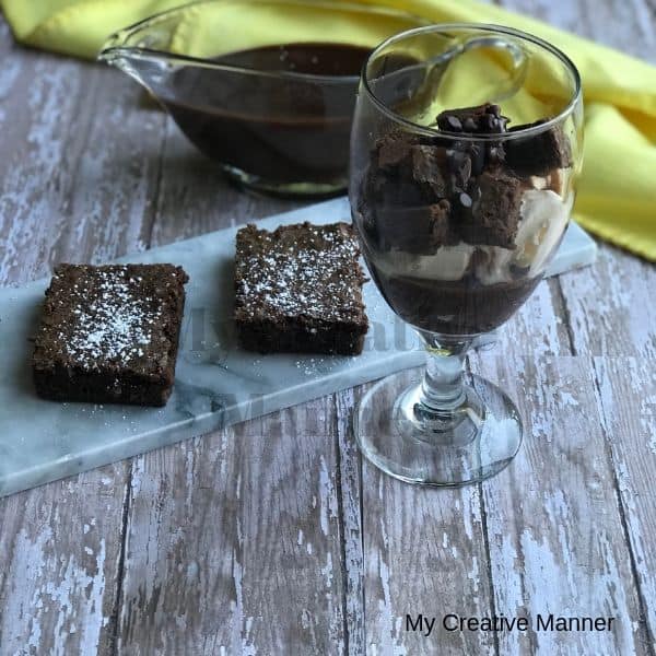 Hot fudge sauce in a gravy boat with a yellow napkin in the background. Three brownies on a plate and a glass that is filled with the Hot Fudge Brownie Recipe.