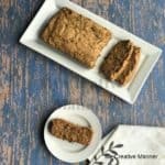 White plate in the background with a loaf of zucchini bread on it. A small white plate with a slice of zucchini bread on it and a white towel in front of it.