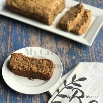 White plate in the background with a loaf of zucchini bread on it. A small white plate with a slice of zucchini bread on it and a white towel in front of it.