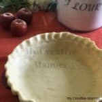 A pie crust that is in a pie dish that has not been baked. There is apples and a flour container in the back ground.