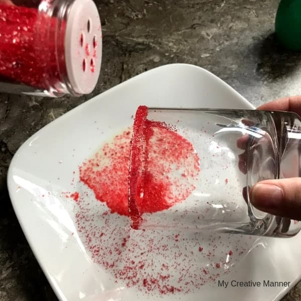 red sugar being sprinkled around a glass that is over a white plate.