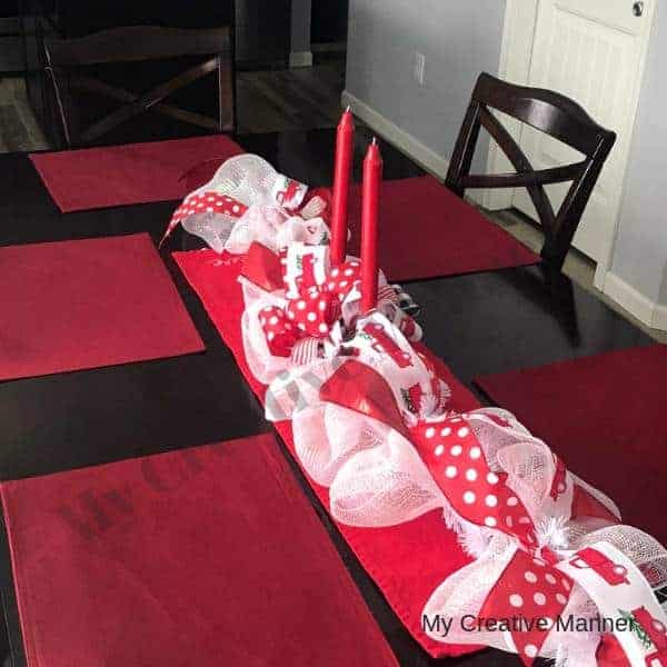 Table with red placemats and a center piece that is made with white mesh and red ribbon. Candle sticks are in the middle of the center piece.