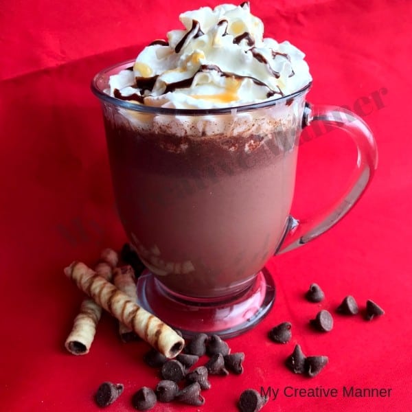 clear glass that has hot cocoa in it with whipped cream on top . , chocolate chips, and cookies lay in from on the cup.