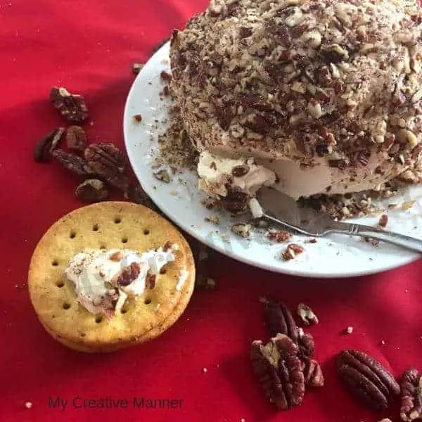 White plate that hold a cheeseball that is covered in pecans with a cheese knife. Pecans and crackers are in front of the plate.