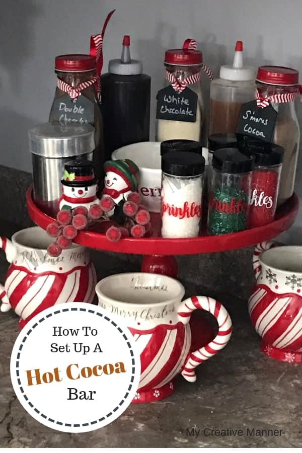 Red cake stand that is filled with containers of hot cocoa, sprinkles and syrup. With three Christmas cups sitting under the cake stand. 