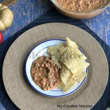 A plate that has beef queso dip and tortilla chips on it that is sitting on a brown plate charger. Behind the chager is a dish that is full of the queso dip.
