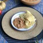 A plate that has beef queso dip and tortilla chips on it that is sitting on a brown plate charger. Behind the chager is a dish that is full of the queso dip.