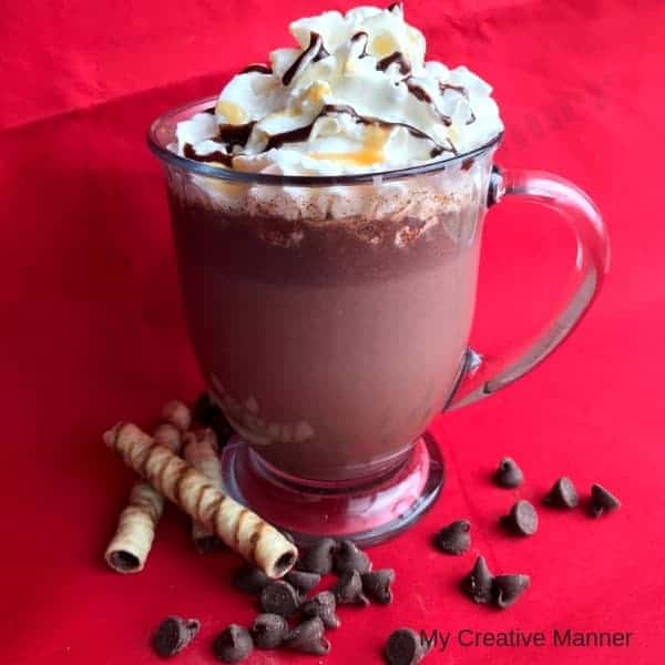 Clear mug with hot cocoa in it that is topped with whipped topping with caramel and chocolate syrup on it. With cookies and chocolate chips in front of the cup. 