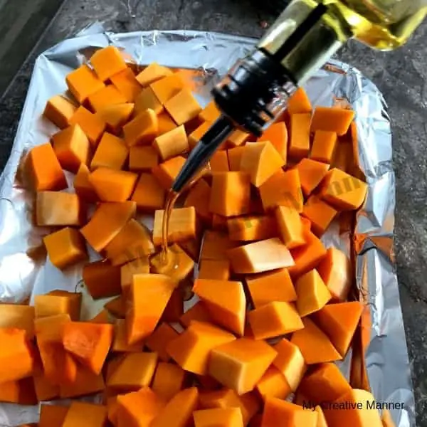 Butternut squash chunks on a cookie sheet that is covered in foil. With a bottle of olive oil being poured on the squash.