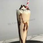 Tall glass filled with a Frozen Mudslide cocktail recipe that is topped with whipped topping and chocolate shavings.