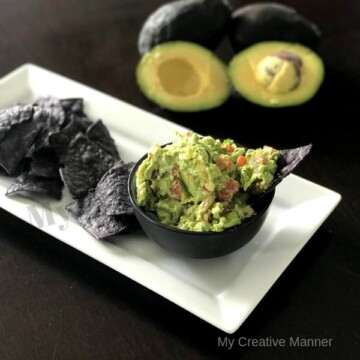 White plate with Blue tortilla chips on it and a small bowl filled with guacamole. With a full and a cut in half avocados in the background.