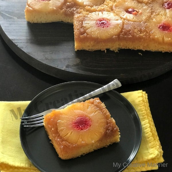 Pineapple upside down cake on a grey plate that is on a yellow napkin.There is a fork on the plate. Behind the plate is a board that has the rest of a 9x13 pineapple upside down cake on it.