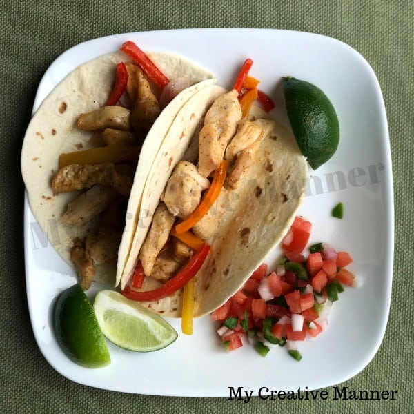 White plate that has two chicken fajitas on it with Pico De Gallo on the side. Along with two slices of lime.