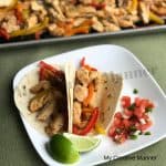 White plate that has two chicken fajitas on it with Pico De Gallo on the side. Along with two slices of lime. Behind the plate is a sheet pan full of chicken and peppers.