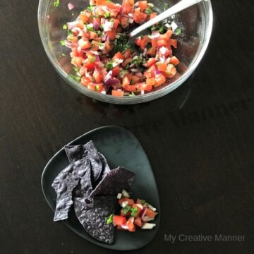 Clear Bowl with Pico De Gallo and a spoon in it. In front of the bow is a grey plate that has Pico De Gallo and Tortilla chips on it.