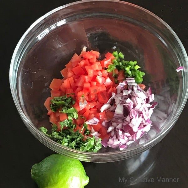 A clear bowl with tomato, onion, cilantro, and jalapño in it. With a lime sitting next to the bowl. 