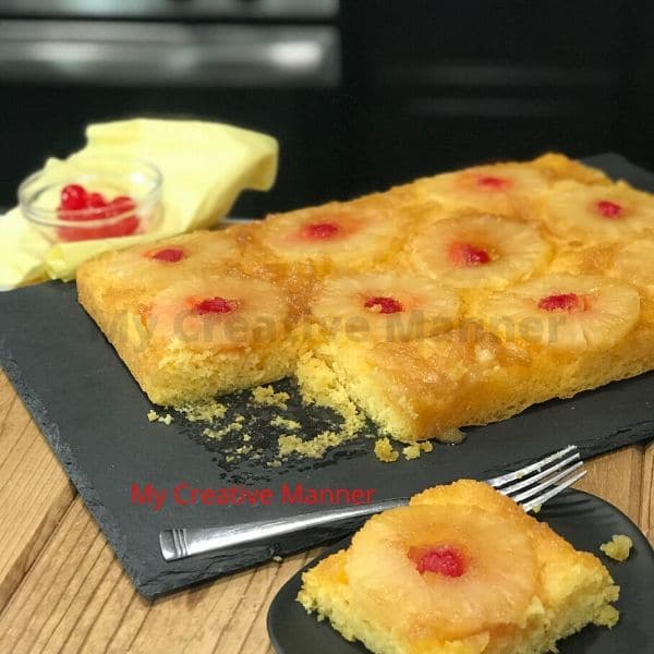 A pineapple upside down cake on a grey platter that is missing a slice of the cake. Behind the platter is a bowl with cherries in it and a yellow napkin.The slice of cake is on another grey plate with a fork on it.