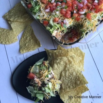 Plate with Seven Layer Taco dip on it and tortilla chips. A baking dish full of the seven layer taco dip and torilla chips laying next to it.