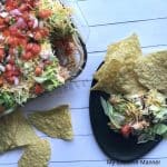 Plate with Seven Layer Taco dip on it and tortilla chips. A baking dish full of the seven layer taco dip and torilla chips laying next to it.