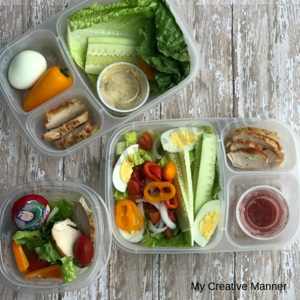 Picture shows three containers that are filled with lunch items. The top left containers has lettuce leaves, cucumber, hummus, chicken breast, mini sweet pepper and a hard boiled egg. The second container has a salad that has tomatoes, sliced hard boiled eggs, and sliced sweet mini pepper in top. There is chicken breast and a container that is filled with salad dressing. The third container has lettuce, mini sweet pepper sliced , whole cherry tomatoes, and a Baby Bell cheese wedge.