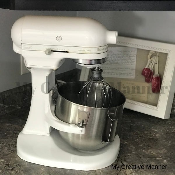 Counter top Kitchen Aid mixer that is in front of a picture that has recipes in it and measuring spoons.
