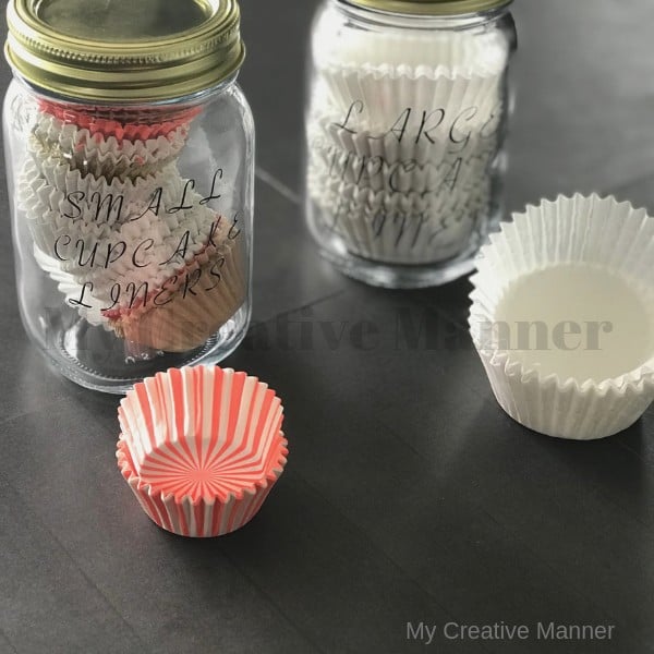Two jars that hold paper cupcake liners. In front of the jars is a stack of each size of the cupcakes liners.