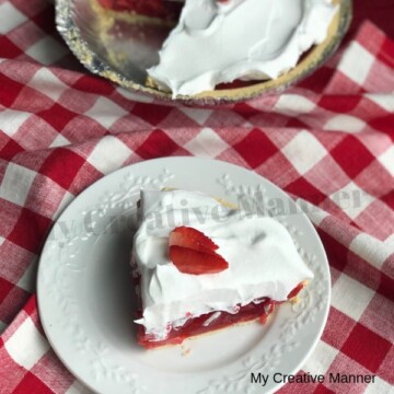 Red and white napkin with a white plate that has a slice of fresh strawberry pie in a shortbread crust that is topped with whipped cream. Behind the plate is the rest of the pie.