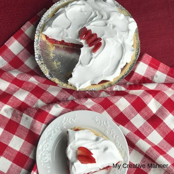 Red and white napkin with a white plate that has a slice of strawberry pie that is topped with whipped cream. Behind the plate is the rest of the pie.