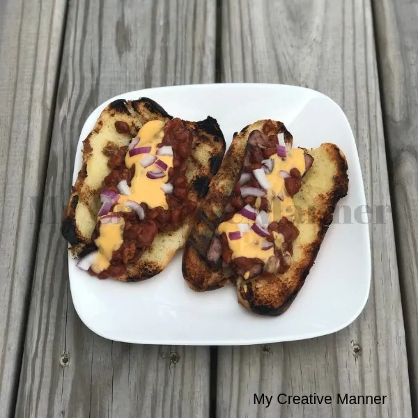 White plate with two Grilled hot Dogs that are in buns and topped with cheese, chili, and red onion.