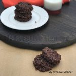 No bake cookies sitting on brown paper and two sitting on a white plate. The whit plate is sitting on a round piece of wood that has a glass of milk on it.