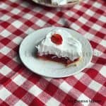 Fresh strawberry jello pie in a shortbread crust that is topped with whipped cream on a white plate that is sitting on a red and white napkin.