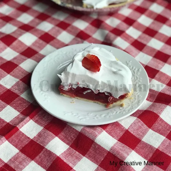 Strawberry pie that is topped with whipped cream on a white plate that is sitting on a red and white napkin.