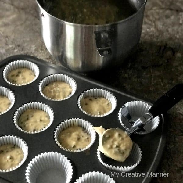 Mixing bowl and in front of that is a muffin tin that has muffin liners and is being filled with batter. There is an ice cream scoop full of the batter filling on of the liners.