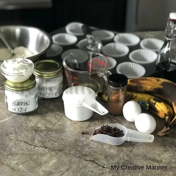 Picture shows measuring cup that is full of sugar, cinnamon jar, jar with baking soda, muffin tin with liners, eggs, and a scoop with mini chocolate chips.