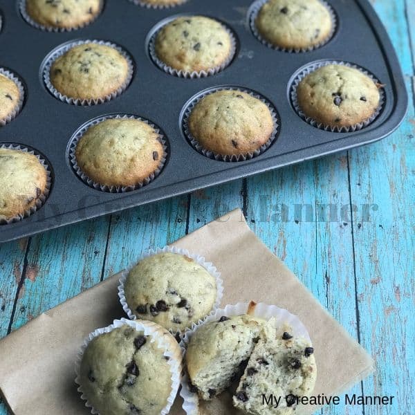Muffin tin with Chocolate Chip Banana Bread Muffins in it. In front of the pan is three muffins on a brown paper. One of the muffins is cut in half.