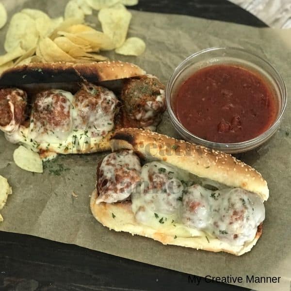 Two meatball sandwiches on brown paper with marinara sauce in a bowl next to it and some potato chips along side the sandwich.