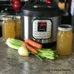 An Instant Pot with three mason jars that are filled with bone broth sitting on a counter top with celery, onion and carrots in front of the jars.