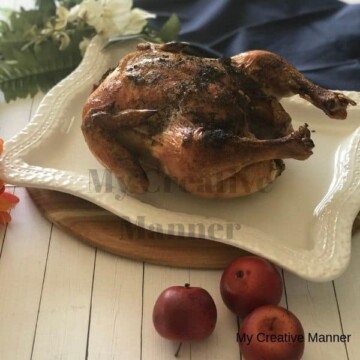 White platter with a whole roasted chicken on it. The platter is on a wood board with a blue napkin behind it. There are flower next to the napkin and apples in front of the platter.